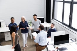 Five co-workers are gathered talking in a bright, modern office. A few are sitting at desks, while others are standing.