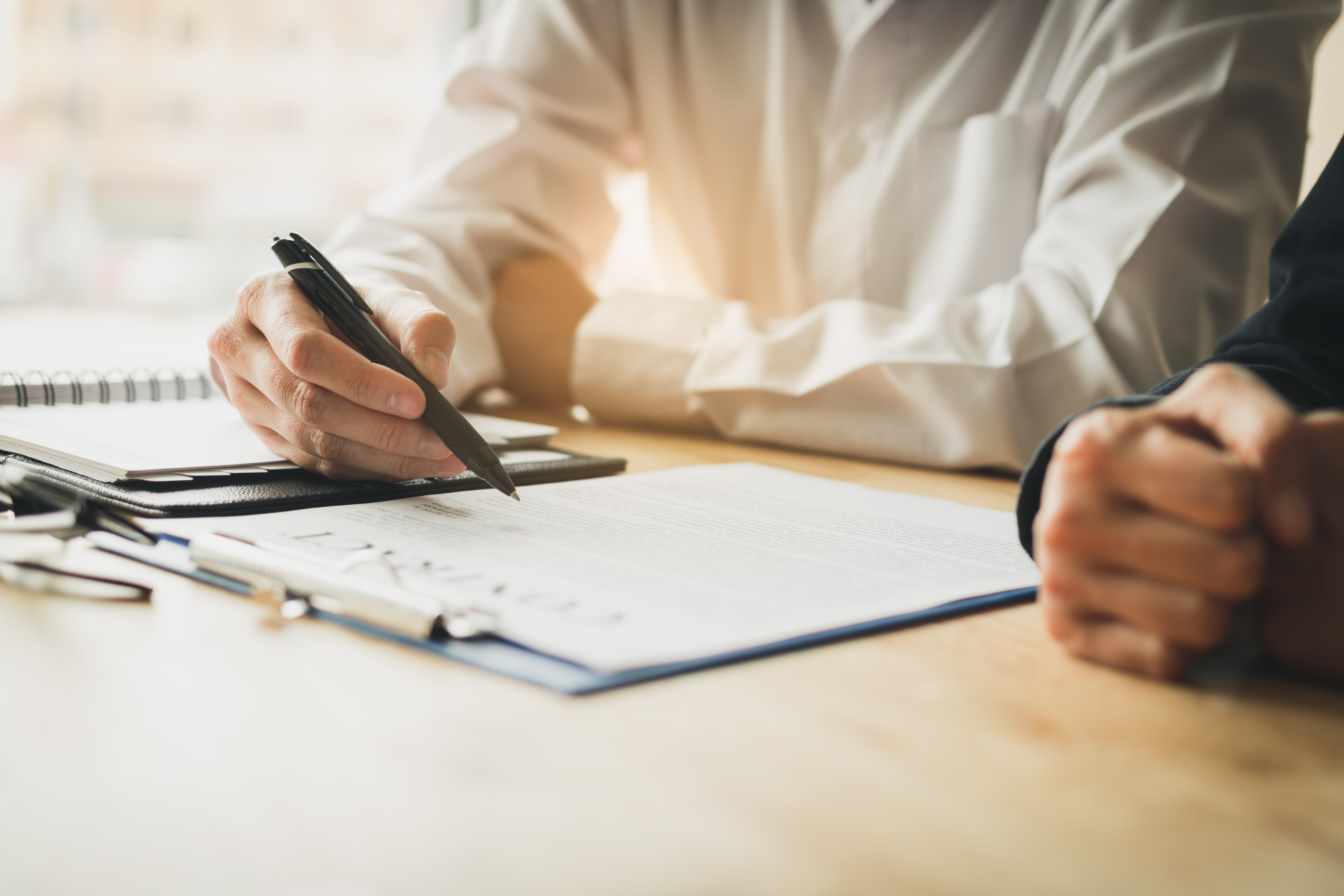 Two people sitting at a wooden desk reviewing and discussing a document on a clipboard. One person is holding a pen and appears to be pointing at or preparing to sign the document, while the other person has their hands clasped together. A notebook and pen are also visible on the desk. The setting suggests a professional or business context.
